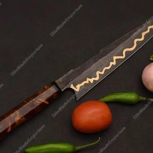 Puede incluir: Cuchillo de chef artesanal con una hoja oscura y un dise&ntilde;o de onda dorada. El mango es marr&oacute;n veteado. El cuchillo se muestra con un tomate, una cebolla y pimientos verdes, lo que sugiere un uso culinario. La hoja mide unos 25 cm.
