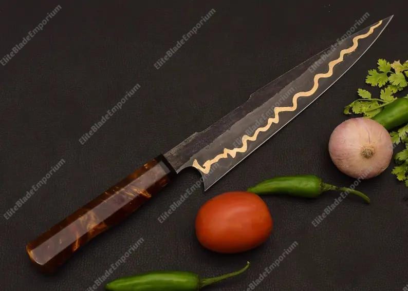 Puede incluir: Cuchillo de chef artesanal con una hoja oscura y un dise&ntilde;o de onda dorada. El mango es marr&oacute;n veteado. El cuchillo se muestra con un tomate, una cebolla y pimientos verdes, lo que sugiere un uso culinario. La hoja mide unos 25 cm.