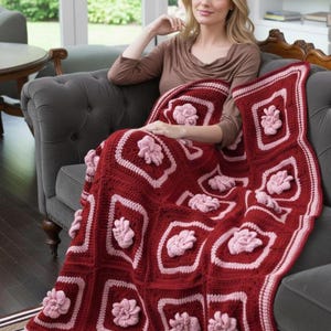 May include: A handcrafted, red crochet blanket with pink floral details. The blanket showcases a repeating pattern of square blocks, each with a raised flower design. The edges are finished with a scalloped border. The blanket is draped over a grey sofa.