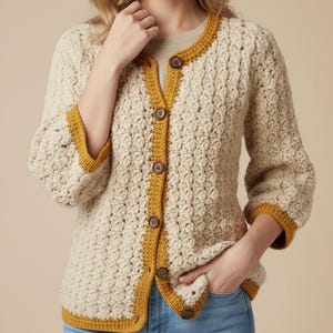 May include: Cream-colored crochet cardigan with a textured stitch pattern. The cardigan features a mustard yellow trim along the edges, sleeves, and neckline. It has three-quarter sleeves and is fastened with brown buttons. The person is wearing blue jeans.