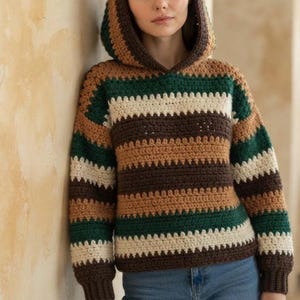 May include: A long-sleeve crochet hoodie featuring horizontal stripes in dark green, cream, and brown. The garment has a hood, ribbed cuffs, and a textured stitch pattern throughout, paired with blue denim jeans for a complete look.