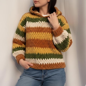Puede incluir: Una sudadera con capucha de ganchillo a rayas, hecha a mano, en tonos marrón, verde, amarillo y crema. El acogedor suéter presenta una capucha y mangas largas con puños acanalados. La sudadera con capucha se combina con unos vaqueros azules.