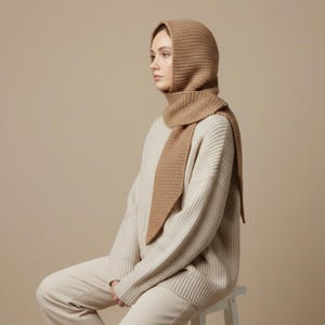 May include: A tan-colored knitted hood and scarf set. The hood covers the head and neck, with a scarf that drapes down the front. The model is wearing a cream-colored sweater and sitting on a white stool. The background is a solid tan color.