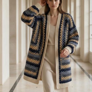 May include: A long-sleeved open-front cardigan featuring a multi-colored crochet pattern in shades of navy blue, charcoal gray, and beige. The garment includes a solid cream-colored ribbed trim along the front opening, cuffs, and bottom hem.