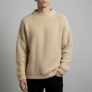 May include: A cream-colored, ribbed knit sweater with a crew neck. The sweater has long sleeves and a relaxed fit. The fabric has a textured appearance. The person in the image is wearing black pants.