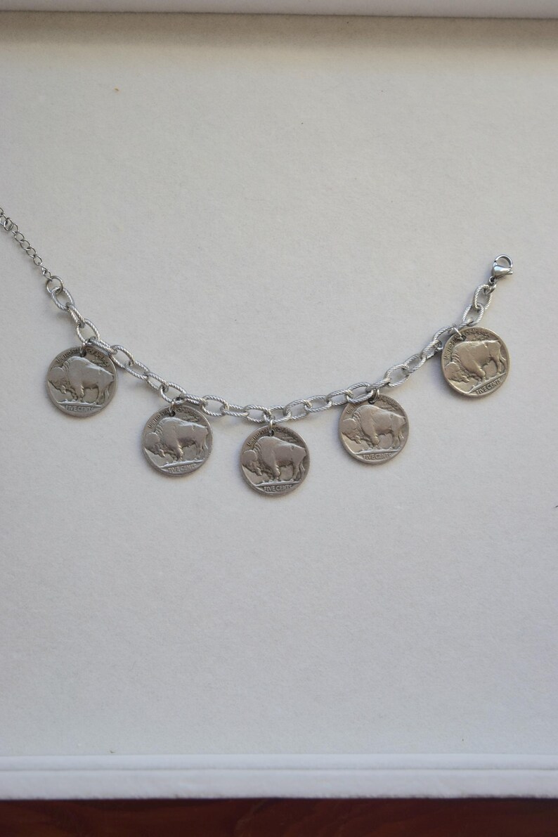 May include: A silver-toned chain bracelet featuring five circular coin charms, each displaying a relief image of an American bison and the text Five Cents. The bracelet includes a delicate link chain and a small metal lobster claw clasp closure.