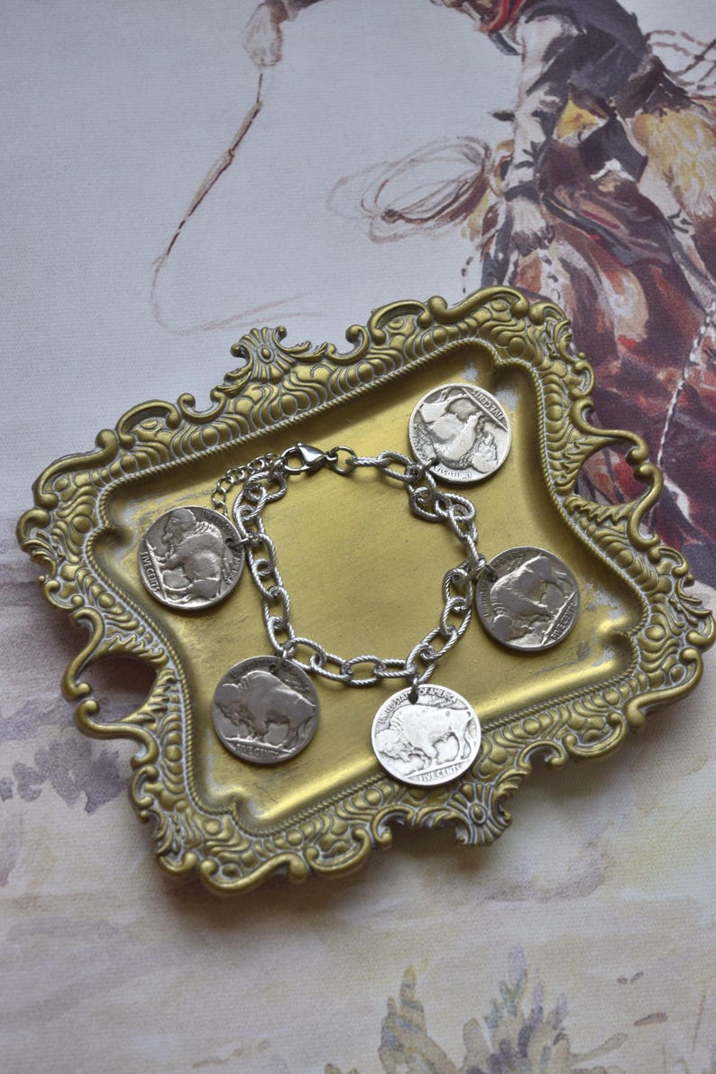 May include: A silver-toned chain link bracelet featuring four circular charms, each depicting a buffalo in profile. The charms are modeled after United States five-cent coins, showing the buffalo design on the reverse side of the metal currency.