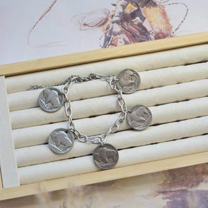 May include: A silver-toned chain link bracelet featuring five circular coin charms, each embossed with the profile of an American bison. The bracelet is constructed with a sturdy metal chain and a secure clasp, showcasing a metallic finish throughout.