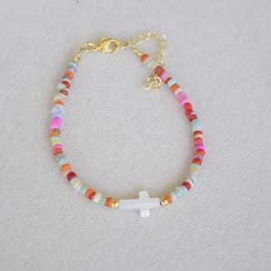 May include: A beaded bracelet featuring a white cross pendant and a gold-colored chain. The bracelet is adorned with small, colorful beads in shades of orange, pink, blue, and green. The clasp and connecting elements are gold-toned.