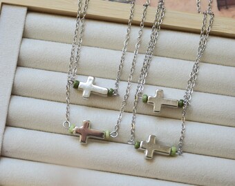 May include: Two silver-toned necklaces featuring a polished cross pendant centered on a delicate chain. Each cross is flanked by small, light green cylindrical beads on both sides, integrated directly into the chain structure for a balanced look.