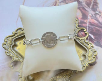 May include: A silver-toned chain bracelet featuring a central circular coin charm depicting a buffalo. The bracelet is constructed with elongated oval links and is displayed wrapped around a small, square, off-white padded cushion.