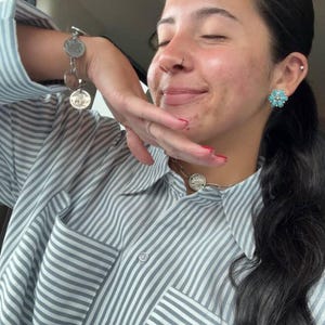 May include: A light blue and white vertical striped button-down shirt with a chest pocket. The jewelry includes a silver-toned coin charm bracelet, a silver-toned chain necklace with a coin pendant, and a flower-shaped turquoise stud earring.