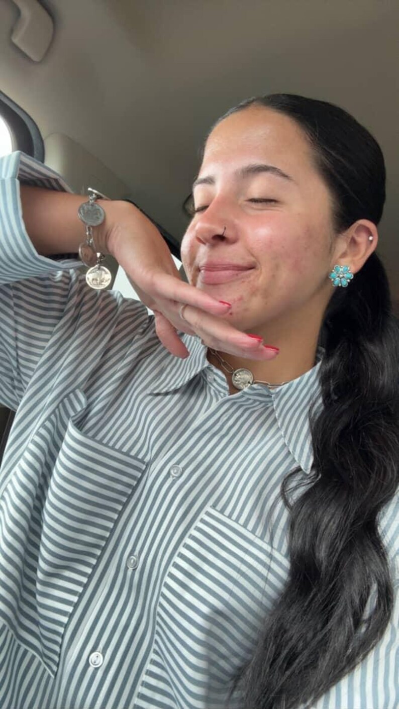 May include: A light blue and white vertical striped button-down shirt with a chest pocket. The jewelry includes a silver-toned coin charm bracelet, a silver-toned chain necklace with a coin pendant, and a flower-shaped turquoise stud earring.