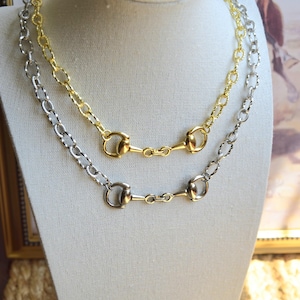 May include: Two necklaces displayed on a mannequin, one in gold-tone metal and one in silver-tone metal. Each necklace features a textured oval link chain connected to a central equestrian-inspired horse bit hardware piece with a polished finish.