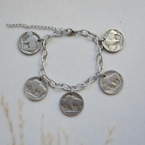 May include: A silver-toned chain link bracelet featuring five circular coin charms. Four of the charms display a buffalo in profile, while one charm shows a Native American figure in profile. The bracelet includes a lobster claw clasp and an extender.