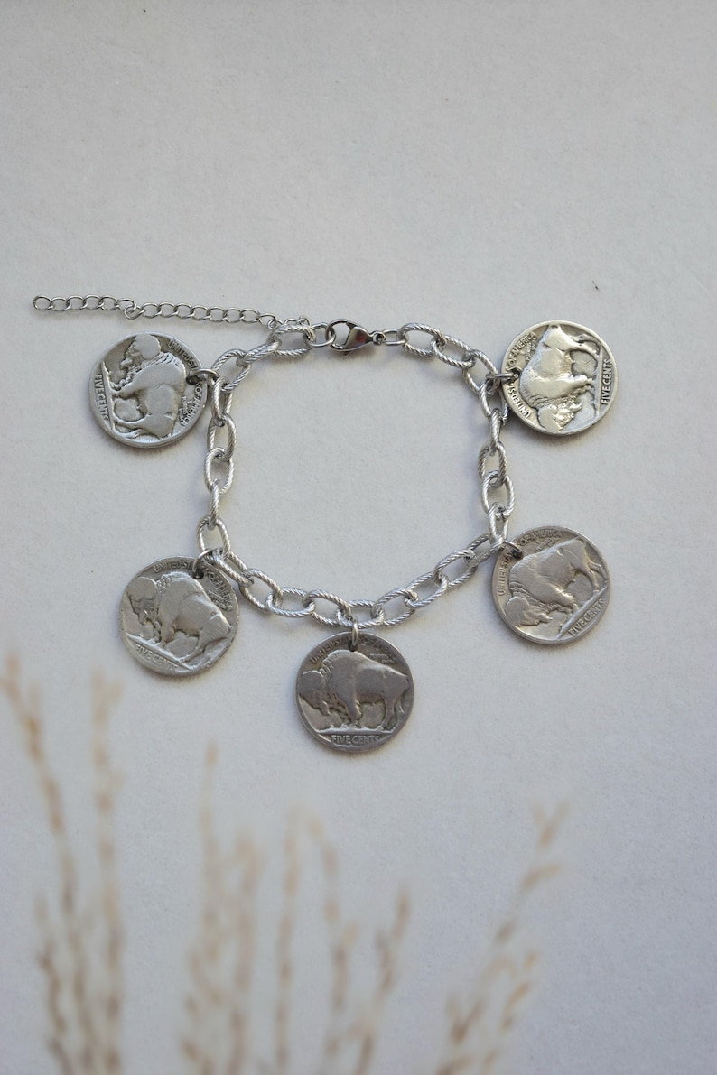 May include: A silver-toned chain link bracelet featuring five circular coin charms. Four of the charms display a buffalo in profile, while one charm shows a Native American figure in profile. The bracelet includes a lobster claw clasp and an extender.