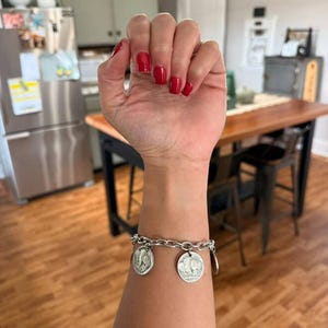May include: A silver-toned chain link bracelet featuring two circular coin charms. Each charm displays a detailed relief of a buffalo on one side and a profile portrait on the other, securely attached to the metal links with small jump rings.