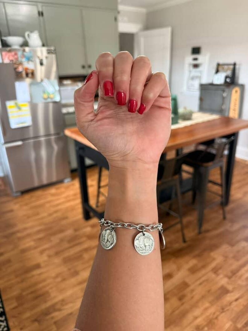 May include: A silver-toned chain link bracelet featuring two circular coin charms. Each charm displays a detailed relief of a buffalo on one side and a profile portrait on the other, securely attached to the metal links with small jump rings.