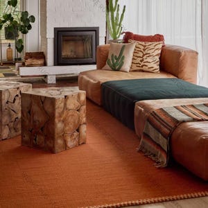 May include: A living room scene featuring a rust-colored rug, a brown leather sectional sofa, and two wooden cube side tables. Decorative pillows with cactus and animal print designs add visual interest. A fireplace and plants complete the cozy, inviting space.