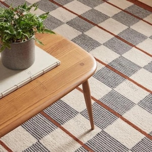 Può includere: Un tavolino in legno con una pianta in vaso e un libro poggia su un tappeto fantasia. Il tappeto presenta uno sfondo color crema con un motivo a griglia di linee nere e marroni. Il tavolo ha una finitura marrone chiaro.