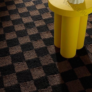 May include: A brown and black checkered rug with a yellow side table. The rug has a repeating pattern of dark brown and black squares. The side table is yellow and has three cylindrical legs. A white decorative bowl sits on top of the table.