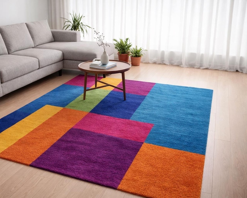 May include: A colorful geometric area rug with a modern design. The rug features large blocks of color in shades of blue, orange, yellow, green, pink, and purple. A gray sofa and a wooden coffee table are in the background.