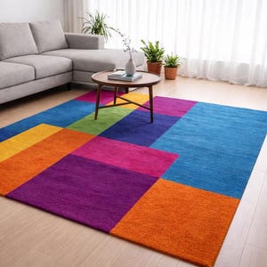 May include: A colorful rectangular area rug with a geometric design. The rug features large blocks of color, including orange, yellow, green, blue, purple, and pink. The rug is placed on a light-colored wooden floor.