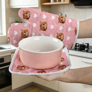 May include: A pink quilted oven mitt and matching square pot holder featuring a repeating pattern of white hearts and orange cat faces. A light pink ceramic bowl rests on the pot holder, which is held by a hand in a kitchen setting.