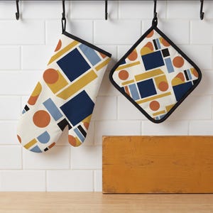 Può includere: Un guanto da forno e una presina quadrata con un motivo geometrico di forme blu navy, giallo senape, azzurro e arancione bruciato su sfondo bianco sporco. Entrambi gli articoli hanno un bordo nero e sono appesi a ganci neri.
