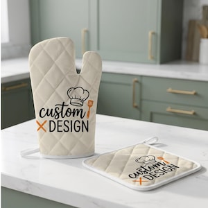 May include: A cream-colored quilted oven mitt and a matching square pot holder. Both items feature a black graphic of a chef hat and a spatula with the text "custom DESIGN" in black, accented by an orange "X" symbol and an orange spatula icon.