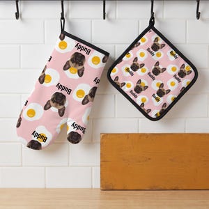 May include: A pink oven mitt and a square pot holder featuring a repeating pattern of fried eggs and the face of a French Bulldog with the name Buddy printed in black text next to each dog face. Both items have black trim and hanging loops.