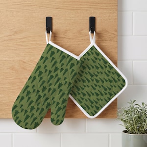 Pode incluir: Uma luva de forno e um descanso de panela quadrado com um padrão repetitivo de pinheiros verde-escuros sobre um fundo verde-oliva claro. Ambos os itens possuem acabamento branco e estão pendurados em ganchos pretos numa superfície de madeira clara.