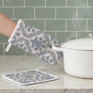 Peut inclure: Un gant de cuisine matelassé et un dessous de plat carré arborant un motif de feuilles botaniques bleues sur fond crème. Le gant possède une boucle en tissu blanc pour le suspendre, et le dessous de plat présente un liseré blanc.