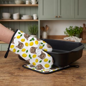 May include: A white oven mitt and matching square pot holder featuring a repeating pattern of brown tabby cat faces, bright yellow lemons, and small green leaves. The oven mitt is shown gripping the edge of a large, rectangular black roasting pan.