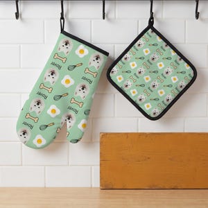 May include: A light green oven mitt and a square pot holder featuring a repeating pattern of white dog faces, fried eggs, yellow whisks, and tan dog bones. The text "Rover" is printed in black next to each dog face on both kitchen items.