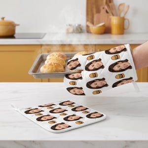 Könnte beinhalten: Ein weißer, gesteppter Ofenhandschuh und ein passender quadratischer Topflappen, beide mit einem sich wiederholenden Muster aus einem Gesicht und kleinen Hamburger-Symbolen. Der Ofenhandschuh hält ein Backblech mit drei Brötchen.