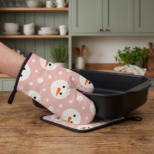 Peut inclure: Un gant de cuisine rose et un dessous de plat carré assorti avec un motif répétitif de visages de bonhommes de neige blancs, nez en carotte orange et yeux noirs, agrémentés de petits flocons de neige blancs, près d'un plat de cuisson noir.