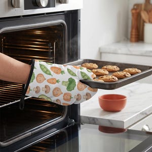 May include: An oven mitt with a white background featuring a repeating pattern of cartoon-style vegetables including carrots, broccoli, mushrooms, peas, and onions, holding a dark metal baking sheet filled with six freshly baked round cookies.