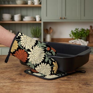 May include: An oven mitt and a matching square pot holder featuring a black background with a repeating floral pattern of large cream, yellow, and burnt orange chrysanthemum flowers accented by muted green leaves, paired with a black baking pan.