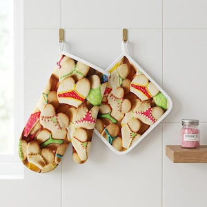 Pode incluir: Uma luva de forno e um descanso de panela quadrado com um padrão repetitivo de biscoitos em forma de coração decorados com glacê colorido em formato de calcinhas de biquíni. Os têxteis de cozinha têm acabamento branco e ganchos dourados.