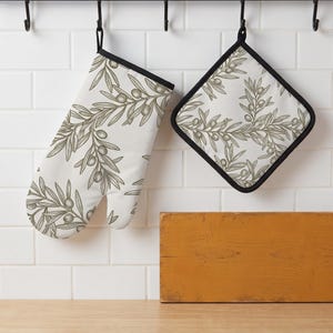May include: An oven glove and a square pot holder featuring a white background with a repeating olive branch pattern in muted green and brown tones. Both kitchen accessories are finished with a solid black fabric trim along the edges.