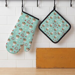 May include: A light teal oven mitt and a square pot holder featuring a repeating pattern of small dog faces wearing white chef hats, red hearts, white paw prints, and white bones. Both items have black fabric trim along the edges.