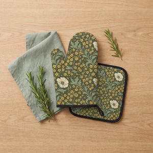 May include: An oven mitt and a square pot holder featuring a dark olive green background with a repeating pattern of small yellow flowers, cream-coloured blossoms, and sage green foliage. The items have black trim and are paired with a sage green cloth.
