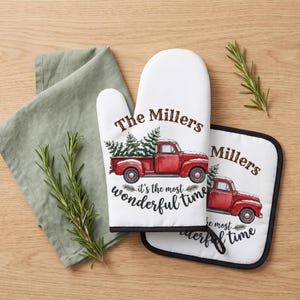 May include: A white oven mitt and a square pot holder featuring a red vintage truck carrying pine trees, with the text The Millers and it's the most wonderful time printed in brown and black script, accompanied by a sage green cloth napkin.