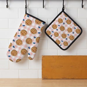Può includere: Un guanto da forno e una presina quadrata con un motivo a quadretti vichy rosa chiaro e bianco. Il tessuto è decorato con una stampa ripetuta di piccole crostate dorate con crosta a griglia e singoli mirtilli blu su tutto il design.