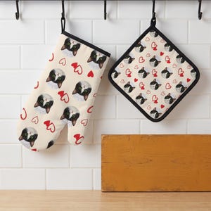 May include: An oven mitt and a square pot holder featuring a repeating pattern of black and white cat faces wearing white chef hats interspersed with small red heart outlines on a light cream-coloured background with black fabric trim edges.