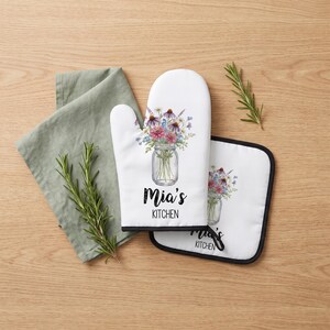 May include: A white oven mitt and a square pot holder, both featuring a watercolor illustration of a mason jar filled with colorful wildflowers. The text "Mia's KITCHEN" is printed in black below the jar on both items. A sage green cloth is included.