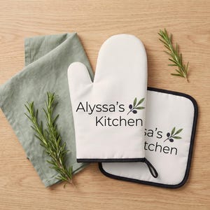 Peut inclure: Un gant de cuisine blanc et une manique carrée assortie avec une bordure noire. Les deux articles portent le texte noir "Alyssa's Kitchen" accompagné d'une petite illustration d'une branche d'olivier avec deux olives et deux feuilles.