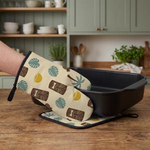 Pode incluir: Uma luva de forno e um descanso de panela quadrado a condizer com fundo creme e um padrão repetitivo de máscaras tiki castanhas, folhas de monstera azul-petróleo, sóis amarelos e palmeiras. A luva é segurada junto a um tabuleiro preto.