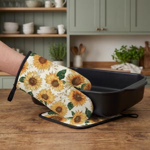 Puede incluir: Una manopla de cocina y un agarrador cuadrado con un patrón repetitivo de girasoles amarillos con hojas verdes sobre un fondo crema. Ambos artículos tienen un borde negro y una trabilla negra, junto a una bandeja de horno rectangular negra.
